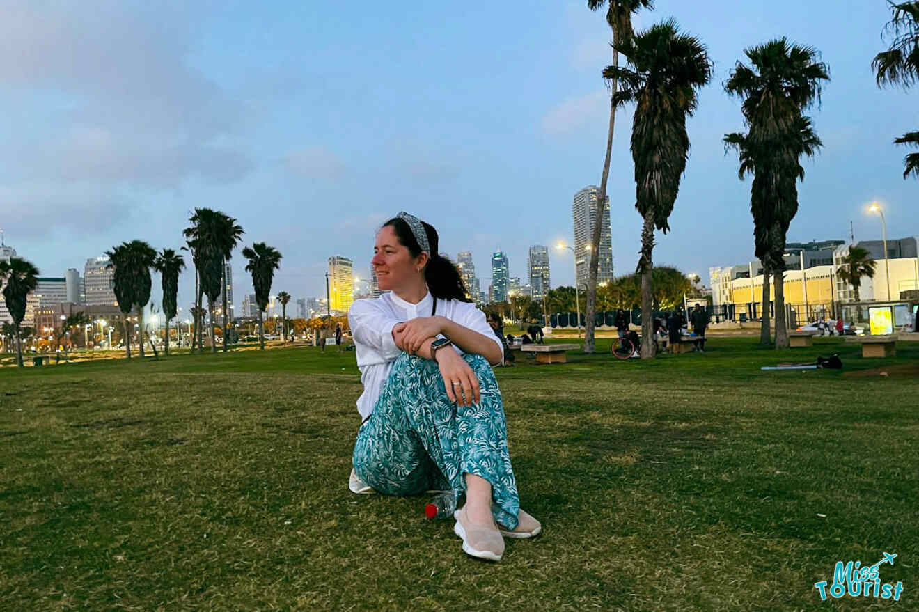 The writer of the post smiling as she kneels on the grass at a park in Tel Aviv during dusk, with the city's skyline in the background