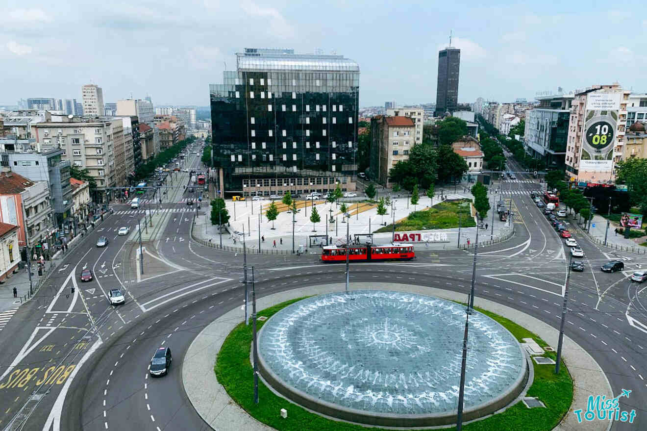An aerial view of a busy Belgrade roundabout with a fountain at its center, depicting urban life and public transport in motion.