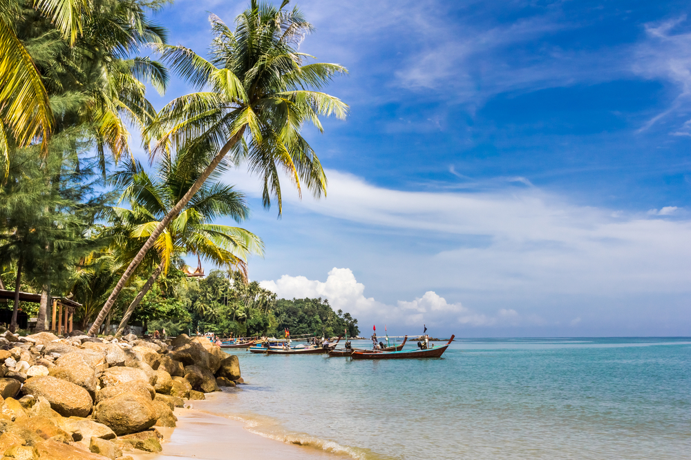 A picturesque view of Bang Tao Beach in Phuket, featuring golden sands with traditional long-tail boats moored in the calm sea, framed by swaying coconut palms under a vast blue sky, encapsulating the serene tropical ambiance of Thailand's coast.





