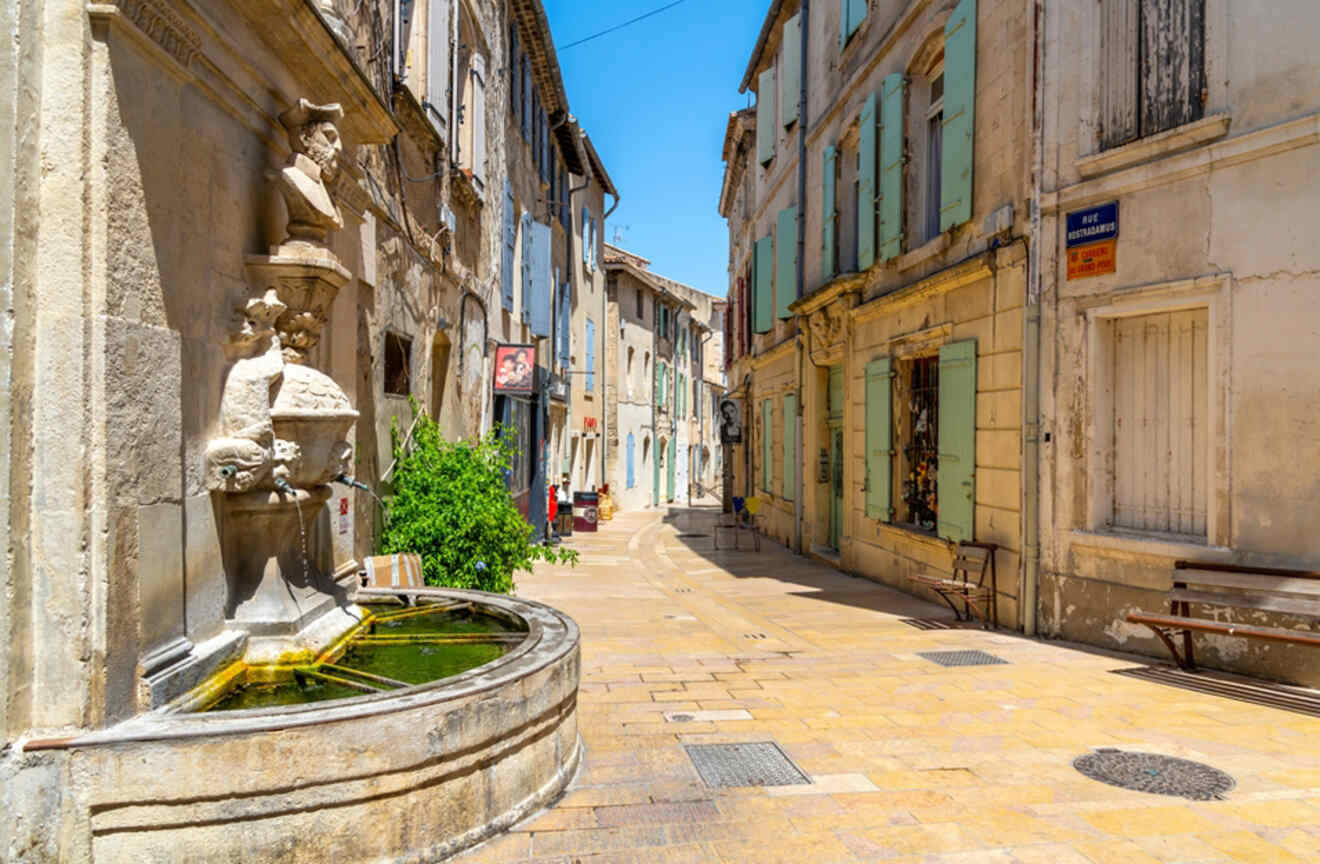 A stone fountain adorned with sculptures on a quiet street in Arles, France, with colorful buildings and a blue street sign for "Rue des Arenes.