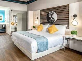 A modern hotel bedroom with a striking wood-panel headboard, white bedding accented with blue and yellow, and stylish bedside lighting.