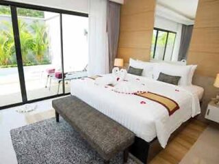 A spacious and elegant hotel bedroom with a large bed adorned with decorative rose petals and a sitting bench, opening out to a pool view through sliding glass doors, epitomizing luxury accommodation.
