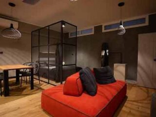 A modern and chic bedroom in Barolo Wine Loft, with a glass partition, a plush red ottoman at the foot of the bed, and stylish hanging lamps providing an intimate atmosphere.
