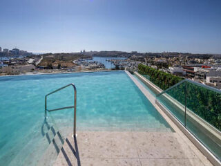 Infinity pool with steps and glass railing overlooking a marina, buildings, and a river under a clear blue sky.
