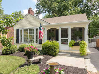 Classic suburban home with beige siding, white trimmed windows, a welcoming front porch, and an American flag