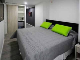 A modern hotel room with a large bed with gray bedding and bright green accent pillows, alongside a kitchenette for convenience.