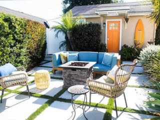 Sunny patio with blue-cushioned furniture, a fire pit, and green plants, in front of a peach house with an orange door.
