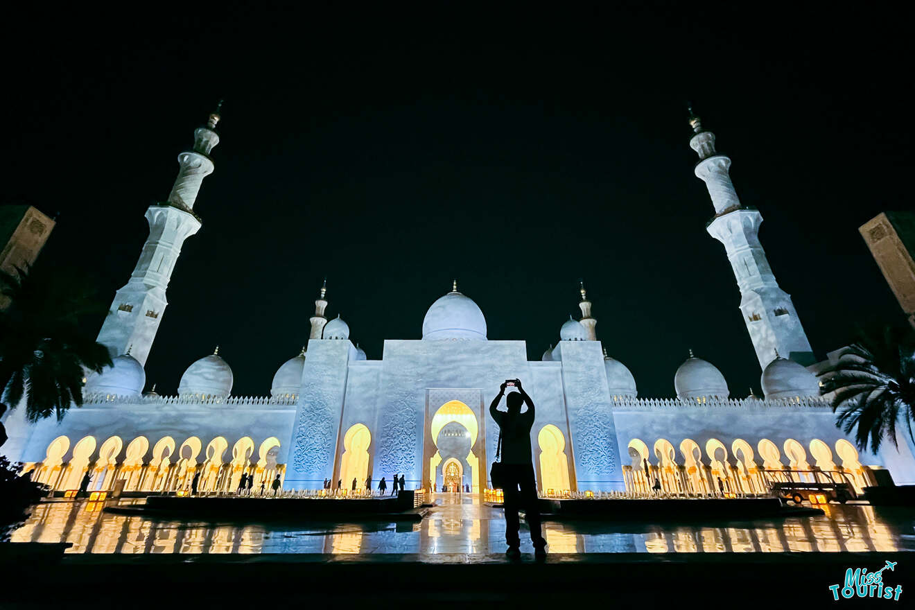 Silhouette of a person making a heart shape with their hands in front of the Sheikh Zayed Grand Mosque in Abu Dhabi, illuminated at night, with the 'Miss Tourist' logo in the corner.