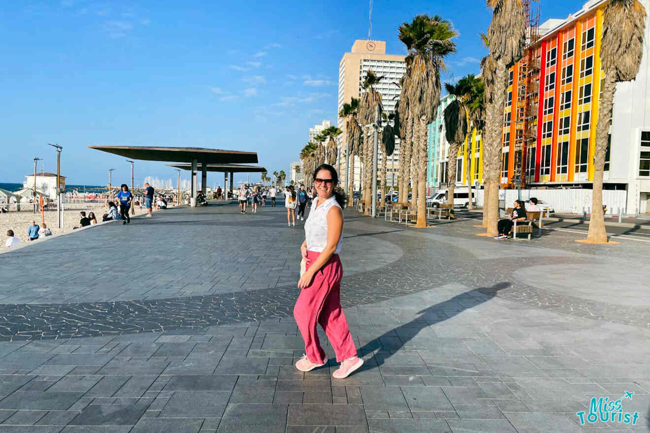 The writer of the post standing on the Tel Aviv promenade, with palm trees and a colorful building façade in the background