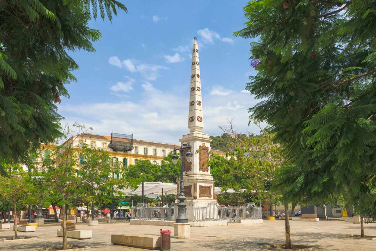 Obelisk monument at La Merced square in Málaga, surrounded by lush trees and a bustling promenade.