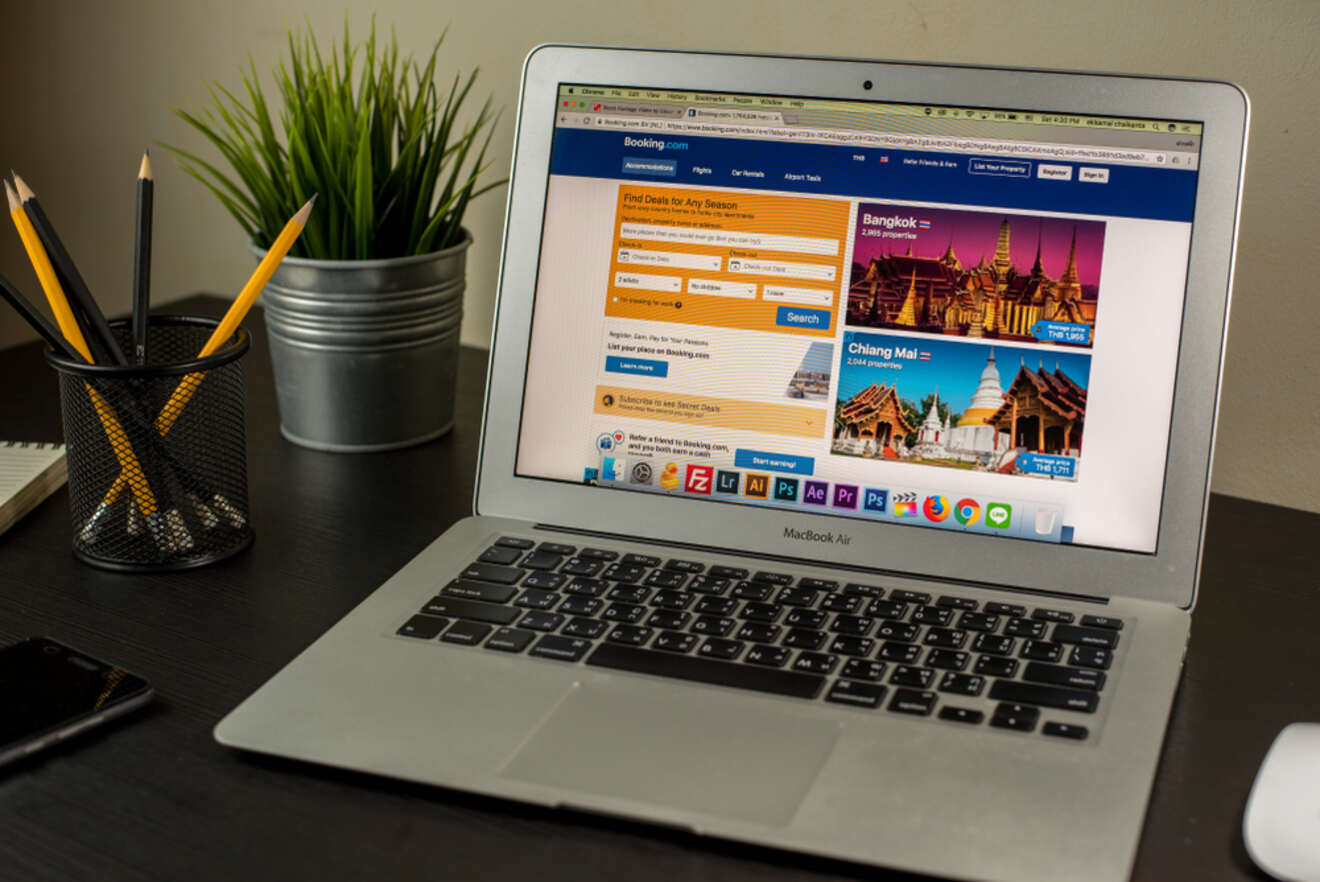 A MacBook Air on a desk displaying Booking.com's homepage with vibrant images of Bangkok and Chiang Mai, offering options to find deals for any season. The workspace is organized with a pencil holder, a potted plant, and a smartphone, creating a professional atmosphere for travel planning