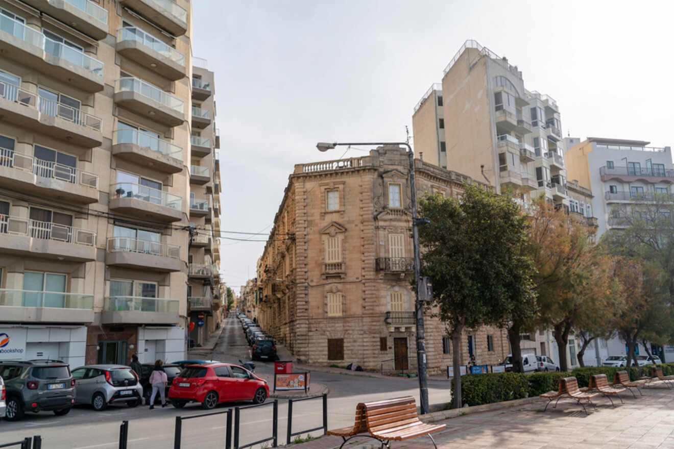A quiet street in Gzira, Malta, showcasing a contrast between the traditional limestone facade of an old corner building and the modern apartment complexes, with parked cars and empty benches on the sidewalk.
