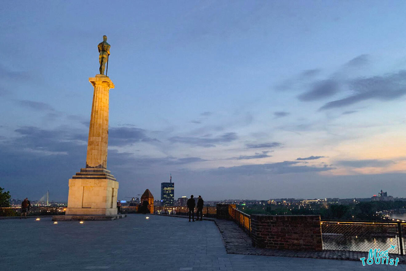 The Victor Monument standing tall against the evening sky at Belgrade Fortress, with the city lights starting to shine in the distance.