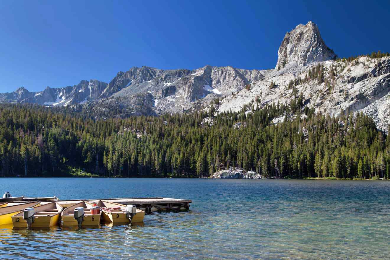 A serene view of Mammoth Lakes with a row of yellow boats docked on a crystal-clear mountain lake, with majestic pine-covered peaks in the background.