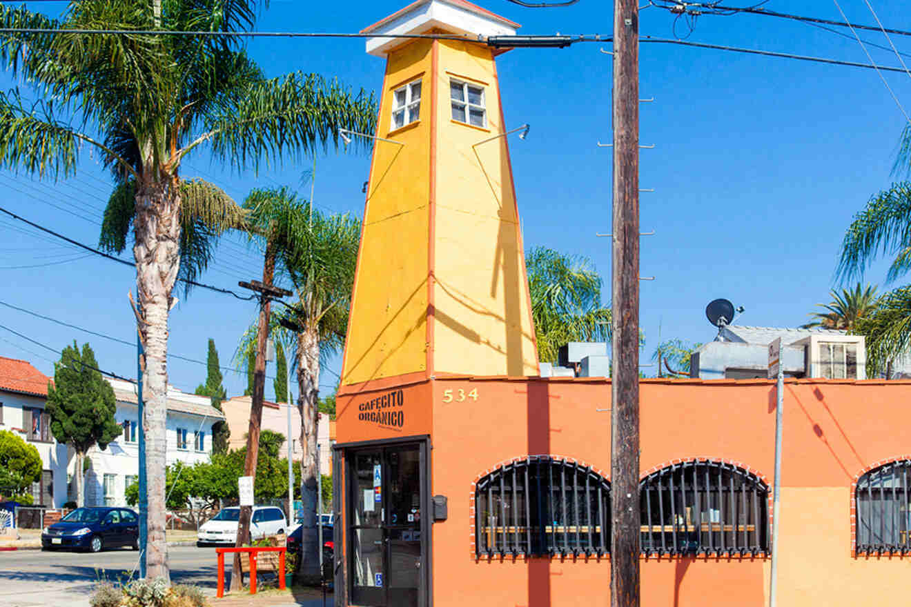 A quirky yellow tower with 'Cafecito Organico' written on the side, nestled in a vibrant neighborhood with palm trees and a clear blue sky.