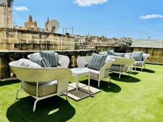 A luxurious hotel rooftop lounge area with plush white seating and striped cushions, set on green artificial turf with historic buildings in the background.