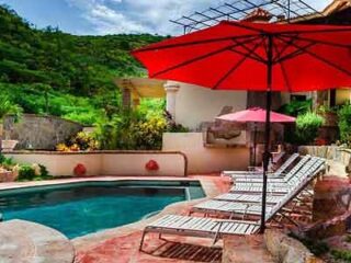 Private poolside area with red umbrellas, lounge chairs, and a lush green hill in the background, creating a serene vacation atmosphere.