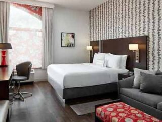 A modern hotel room with a king bed, patterned accent wall, gray sofa, and pops of red from the pillows and lampshade, creating a stylish and comfortable space.