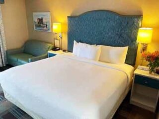 A bright hotel room with a plush king bed, teal armchair, and vibrant bedside flowers, presenting a fresh and inviting atmosphere for guests.