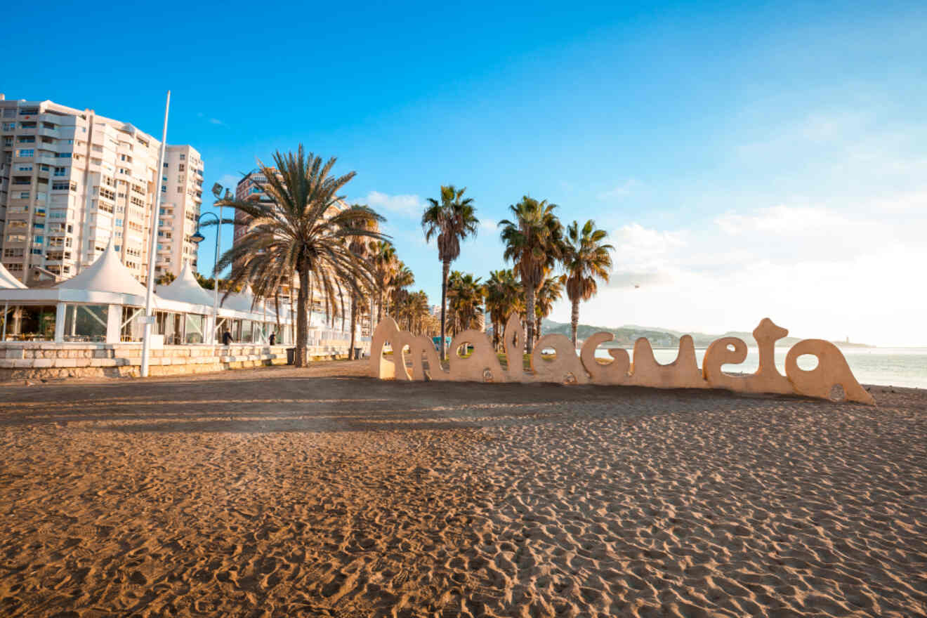 Sandy beachfront of La Malagueta with 'Malagueta' sculpture, palm trees, and beachside buildings under a clear sky