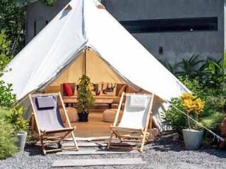 Outdoor glamping scene with a spacious canvas tent open to a cozy interior, flanked by wooden deck chairs, set within a lush garden for a rustic-chic outdoor living experience.
