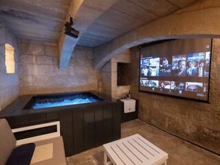A small room with stone walls features a hot tub, a sofa, a white table, and a large projector screen displaying a streaming service menu.