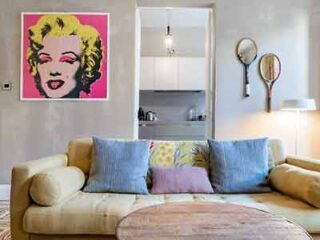 A cozy living room with a modern pop art Marilyn Monroe portrait, a curved sofa with mixed pattern cushions, and vintage tennis rackets on the wall.