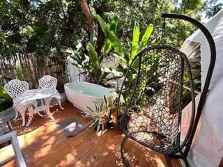 Quaint outdoor bathing area with a freestanding bathtub surrounded by tropical plants, open-air shower, and a black hanging egg chair, offering a unique blend of nature and comfort.