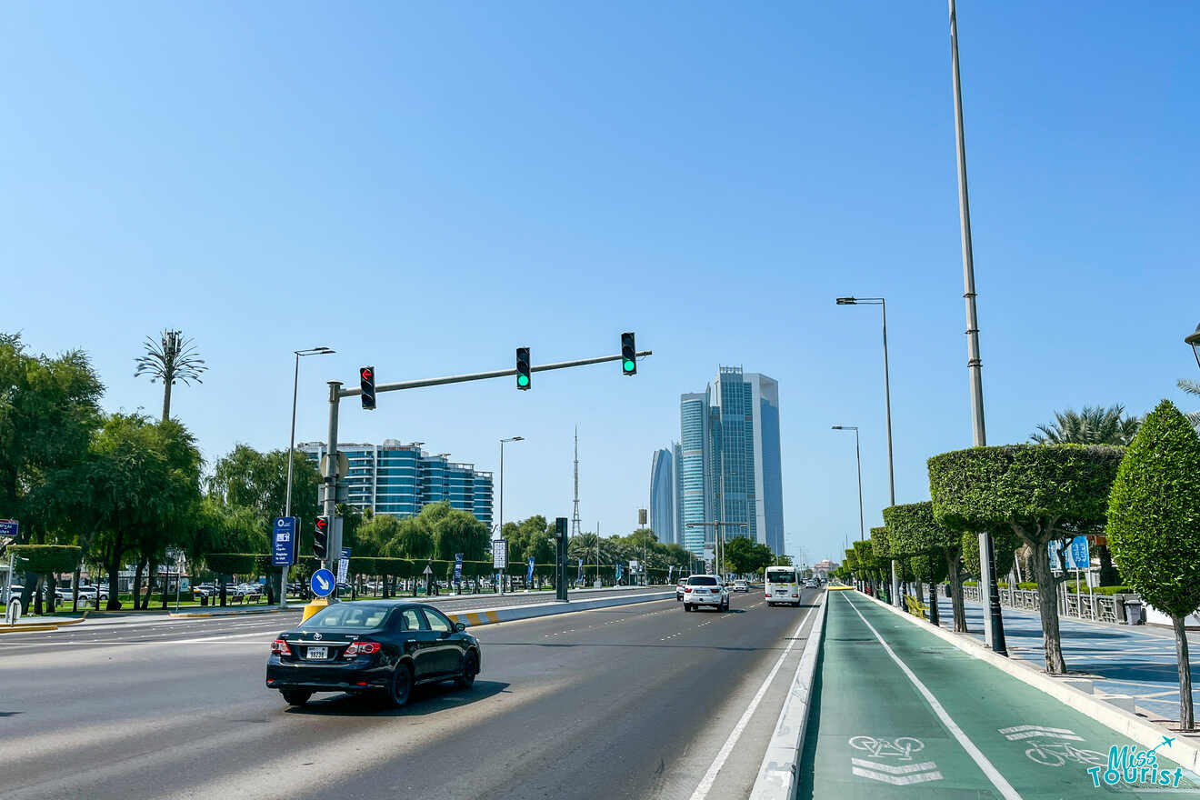 View of a bustling street in Al Khalidiya, Abu Dhabi, with modern skyscrapers in the distance, traffic lights on green, and a dedicated bicycle lane, complete with the 'Miss Tourist' watermark.