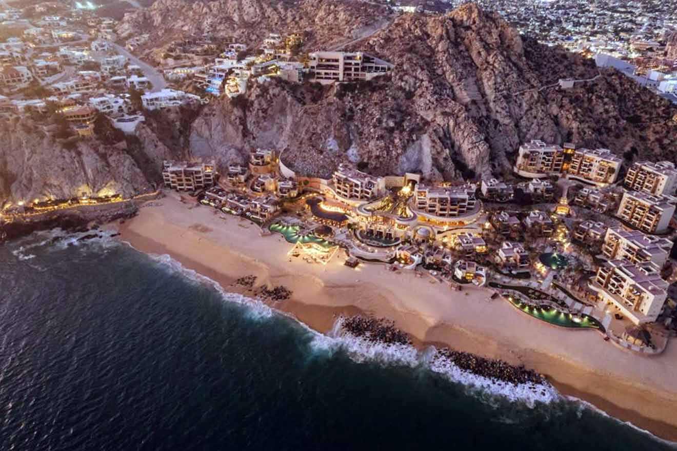 Aerial view of a beachside resort at twilight, with illuminated pools and buildings nestled between the beachfront and rugged cliffs.