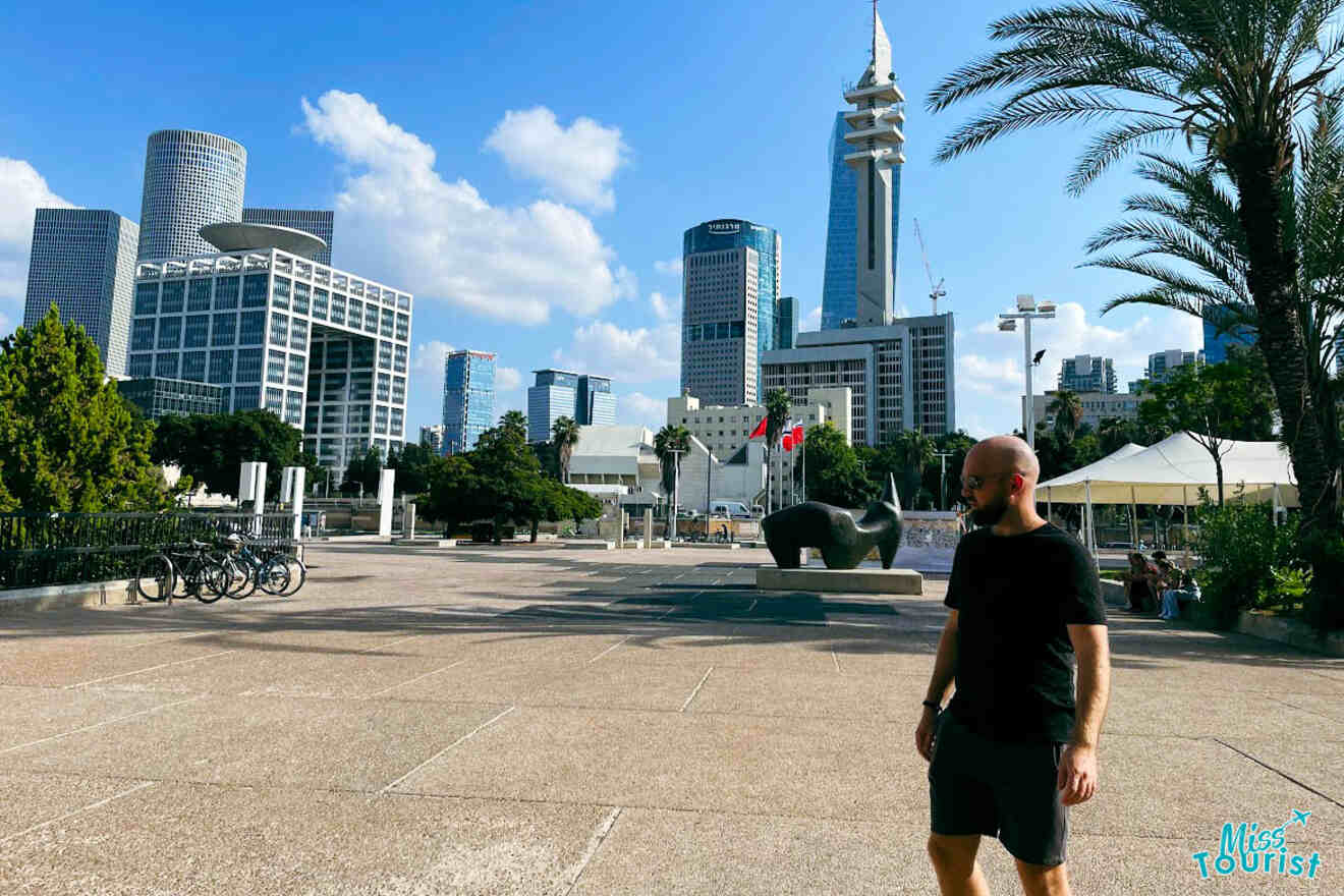 The writer's partner walking past the modern Tel Aviv Museum of Art, with its distinctive architecture and surrounding skyscrapers under a clear blue sky