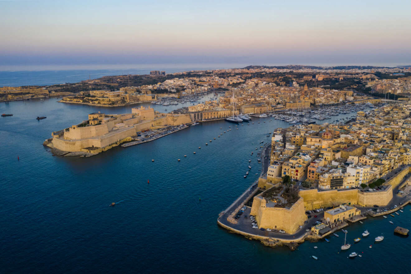 Aerial view of the Grand Harbour in Valletta, Malta at dusk, with the historic Fort Saint Angelo and densely packed buildings along the coastline, and numerous boats and yachts dotting the tranquil harbor waters.