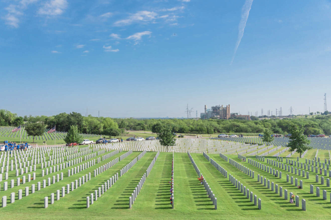 A serene military cemetery in Fort Worth, with uniform white headstones neatly arranged in rows, under a clear blue sky. The meticulously maintained green lawn stretches towards a distant industrial backdrop, symbolizing a peaceful resting place for veterans
