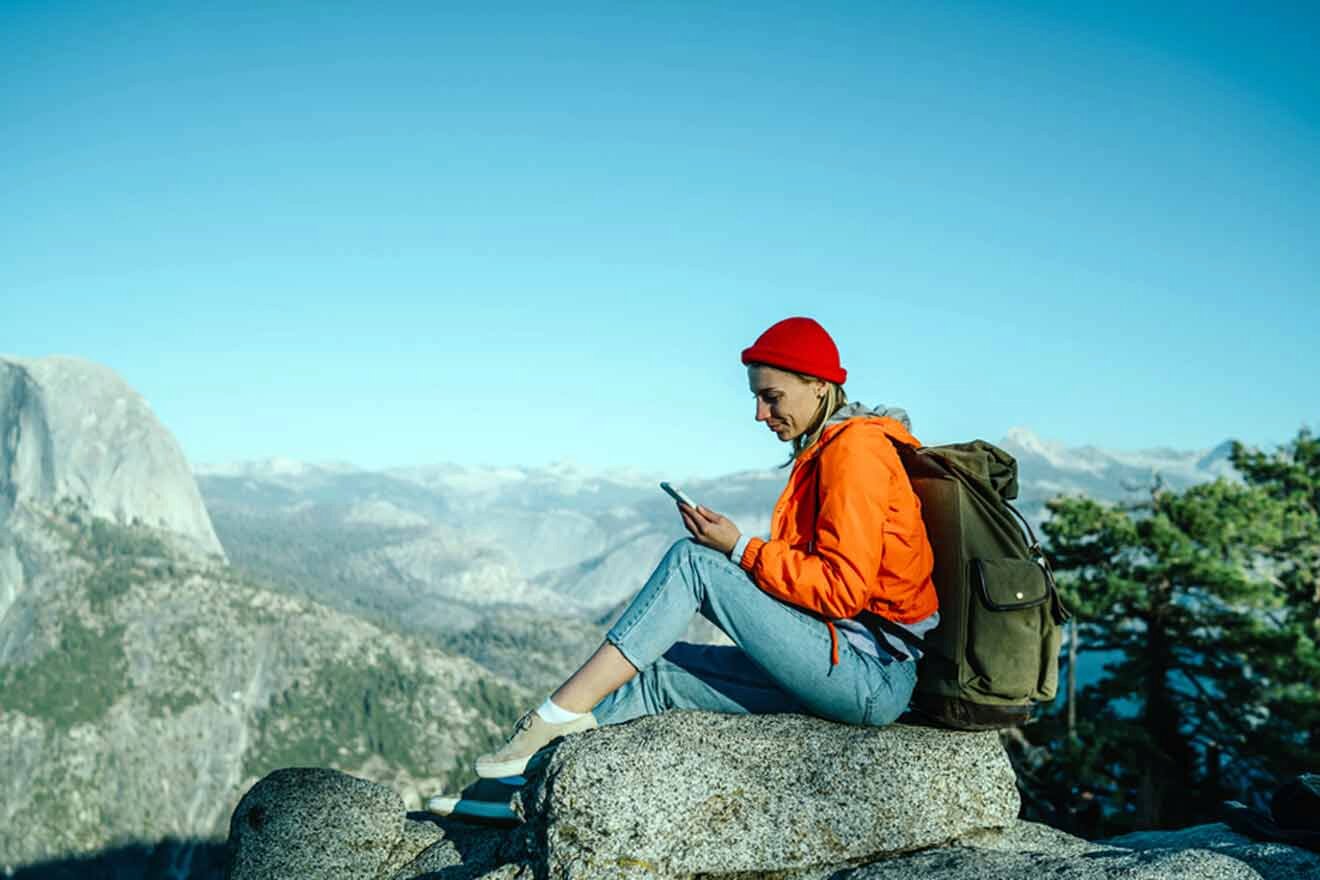 Woman in a red beanie and orange jacket sitting on a rock using her smartphone with a panoramic view of Yosemite's cliffs in the background.