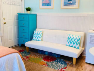 A cozy corner of the Sunny Daze Tiny Cottage featuring a white futon with teal chevron cushions, a vibrant floral area rug, and a turquoise dresser against a pastel blue wall adorned with beach-themed art.