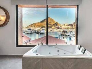 A luxury bathroom with a large jacuzzi tub facing a window that offers a panoramic view of a marina and surrounding landscape.