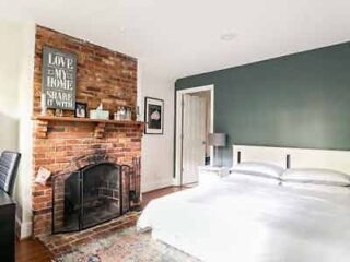 A charming bedroom with exposed brickwork, a fireplace, and wall art reading "LOVE MAKES A HOUSE A HOME," paired with a crisp white bedspread and dark green accent wall.