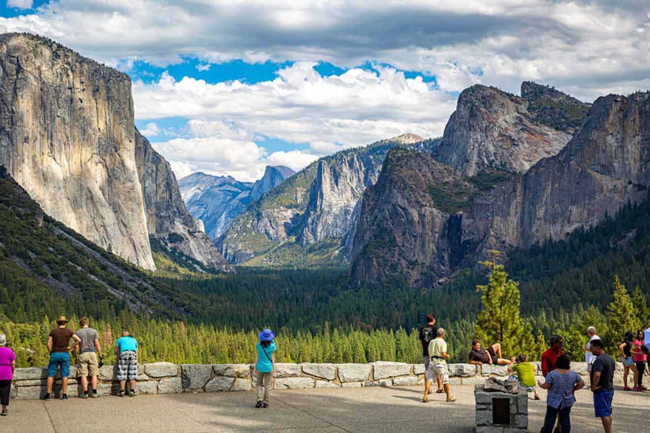 Tourists at an overlook admiring the iconic view of El Capitan and Half Dome in Yosemite Valley, with lush greenery and a clear blue sky.