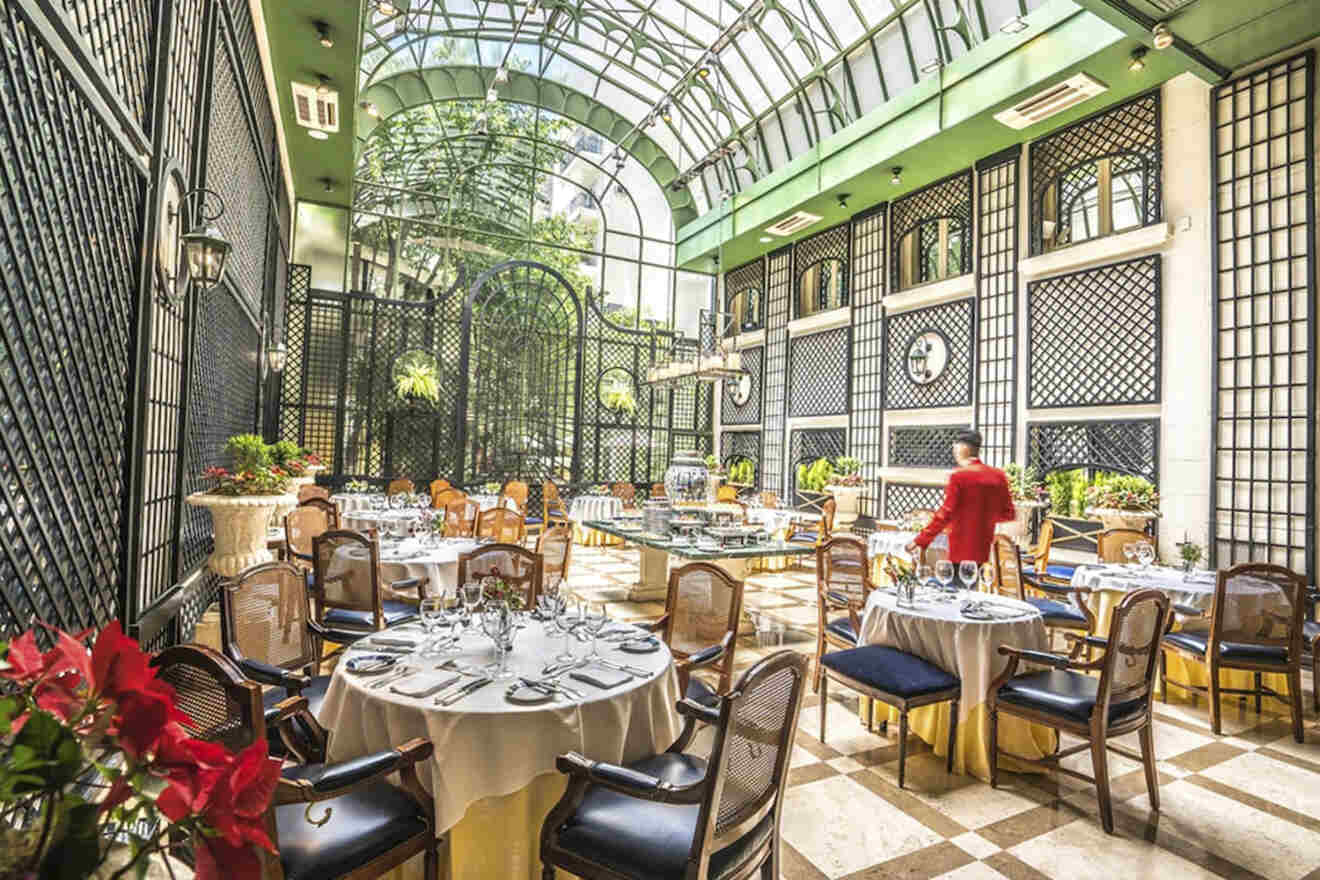 The elegant interior of a restaurant with natural lighting, featuring a glass ceiling, sophisticated décor, and a server in red attire