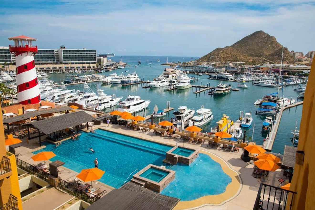 Overlooking a marina and pool area with bright orange umbrellas, a striped lighthouse structure, and boats docked under a clear blue sky.