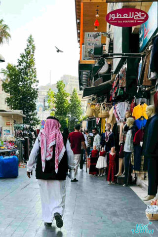 A man in traditional Jordanian attire walking through a lively street market in Amman, with storefronts displaying a variety of goods
