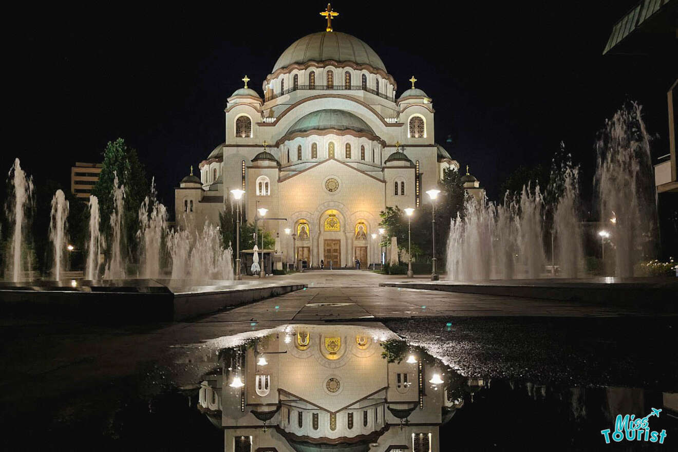 The Saint Sava Church in Belgrade illuminated at night, featuring grand architecture with water fountains in the foreground reflecting the structure