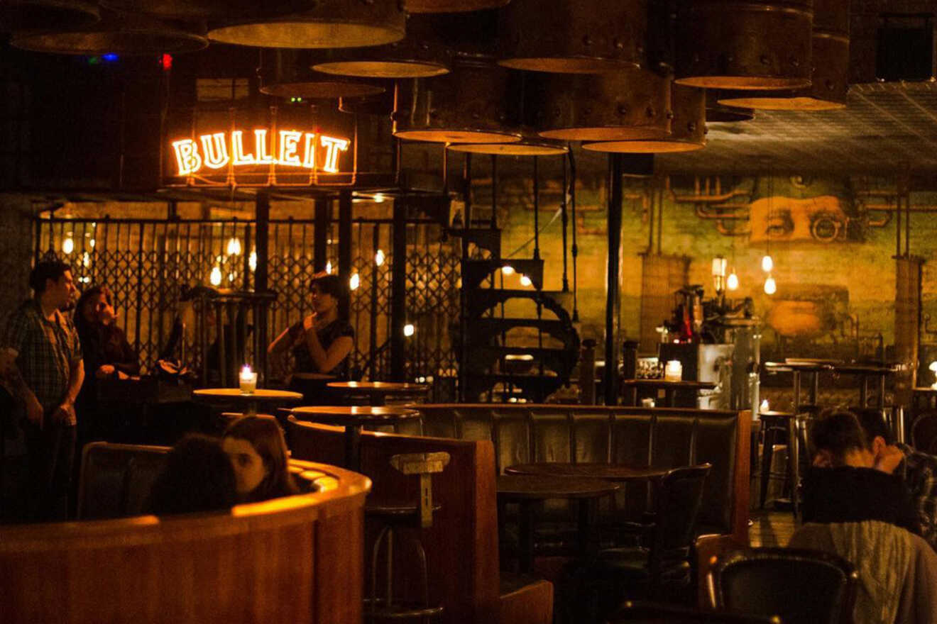 An atmospheric view of Victoria Brown Bar, showcasing a neon 'Bullitt' sign illuminating the dark ambiance, patrons mingling in the intimate seating area with a distinctive industrial chic decor.