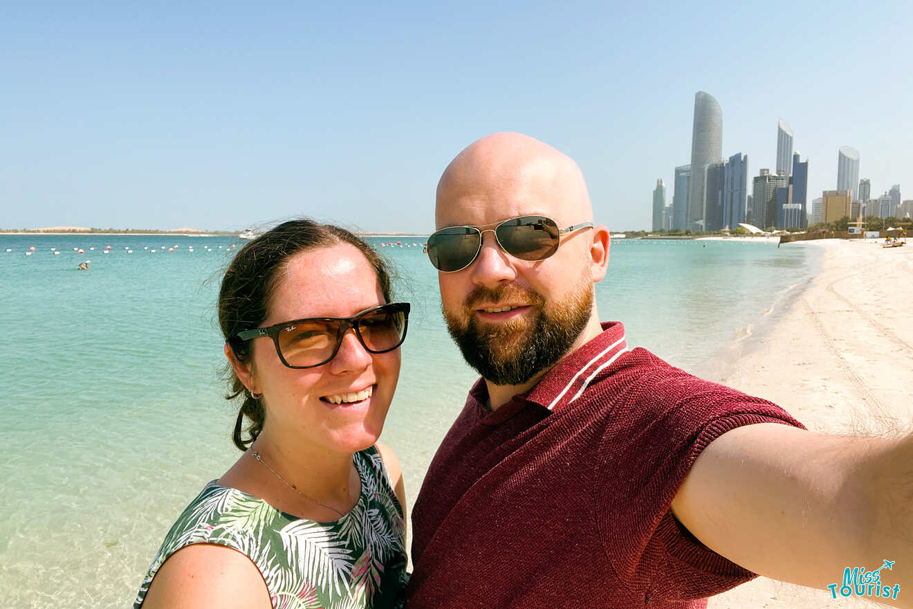 Writer of the post with her husband takes a sunny beachfront selfie with the Abu Dhabi skyline in the background, the clear turquoise waters of the Corniche Beach beside them, and the 'Miss Tourist' logo visible.