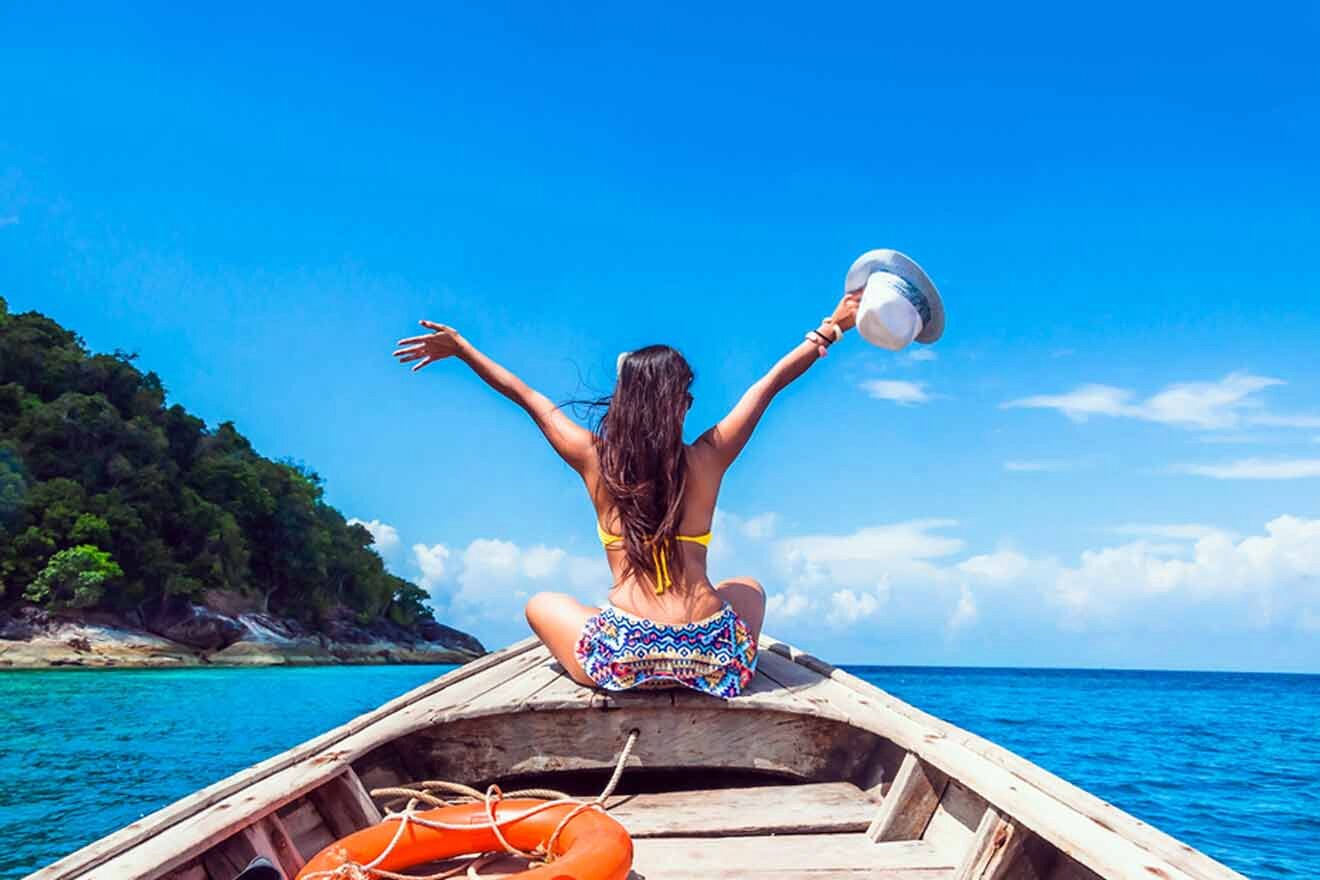 A traveler celebrating freedom and adventure on the bow of a traditional boat, arms outstretched towards the sky, with a clear blue sea and rocky island landscape ahead.