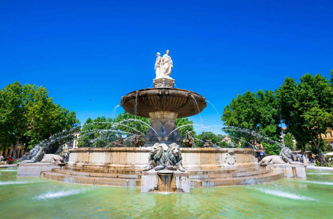 La Rotonde fountain in Aix-en-Provence, France, with statues and lions spouting water, surrounded by lush trees against a clear blue sky