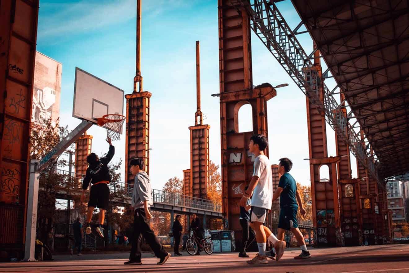 An urban outdoor basketball court in Parco Dora San Donato, bustling with players and onlookers under a structure reminiscent of Turin's industrial heritage.