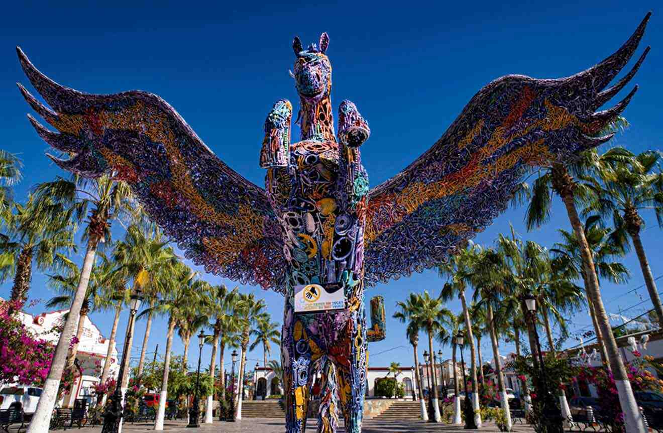 Sculpture of a winged horse, Pegasus, made of colorful mosaic tiles, standing in a plaza surrounded by palm trees under a clear sky.