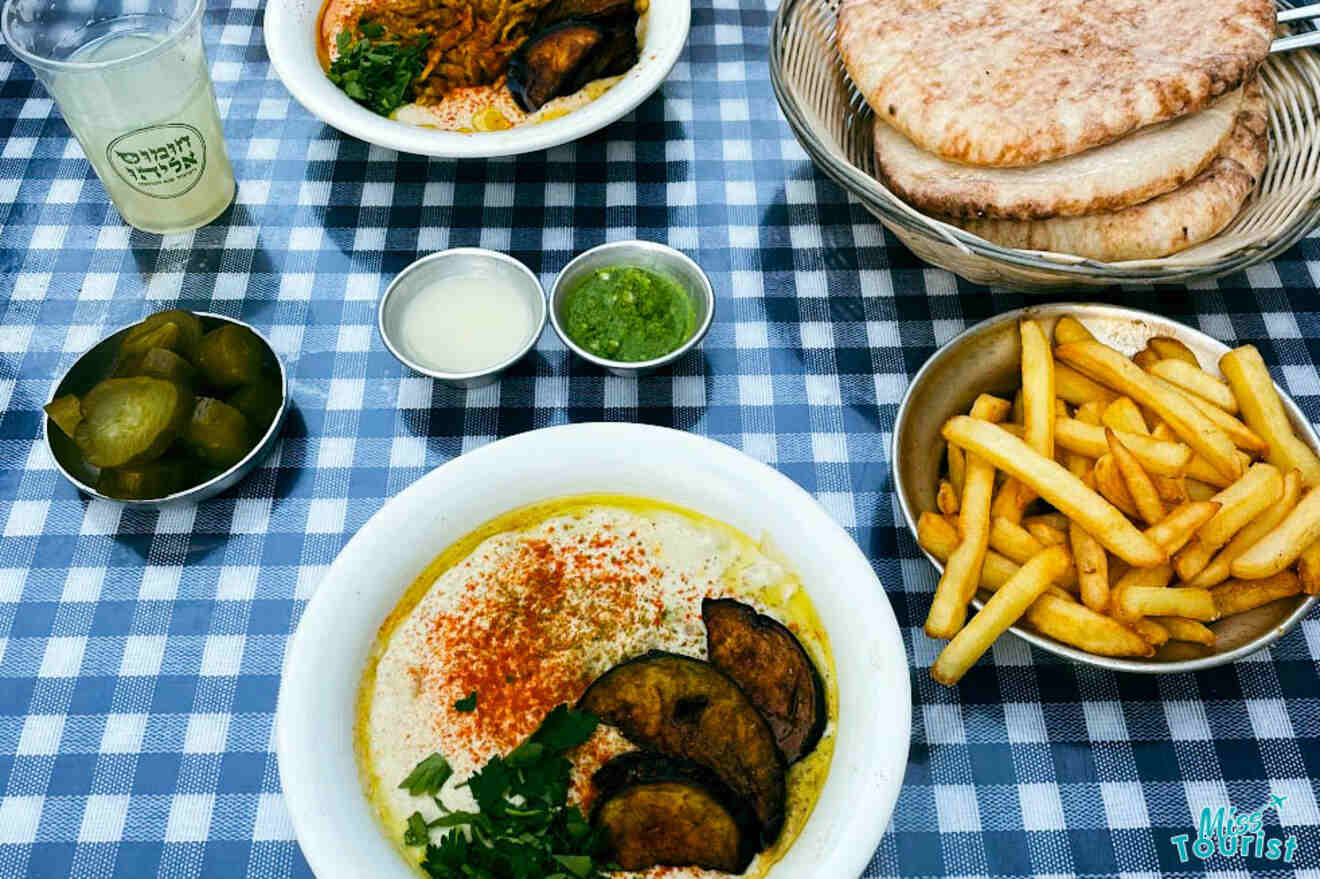 A table with traditional Israeli dishes including hummus with toppings, pita bread, french fries, and pickles, served with a glass of lemonade