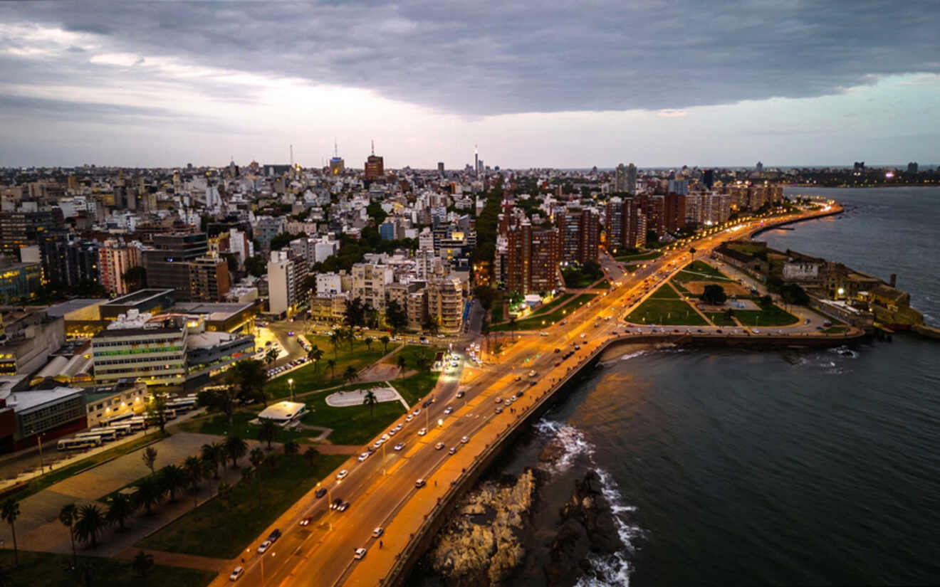 Aerial view of Montevideo at dusk, showcasing the city lights beginning to illuminate the evening, with the Rambla coastline curving along the shore, cars streaming by, and the urban landscape stretching into the distance.